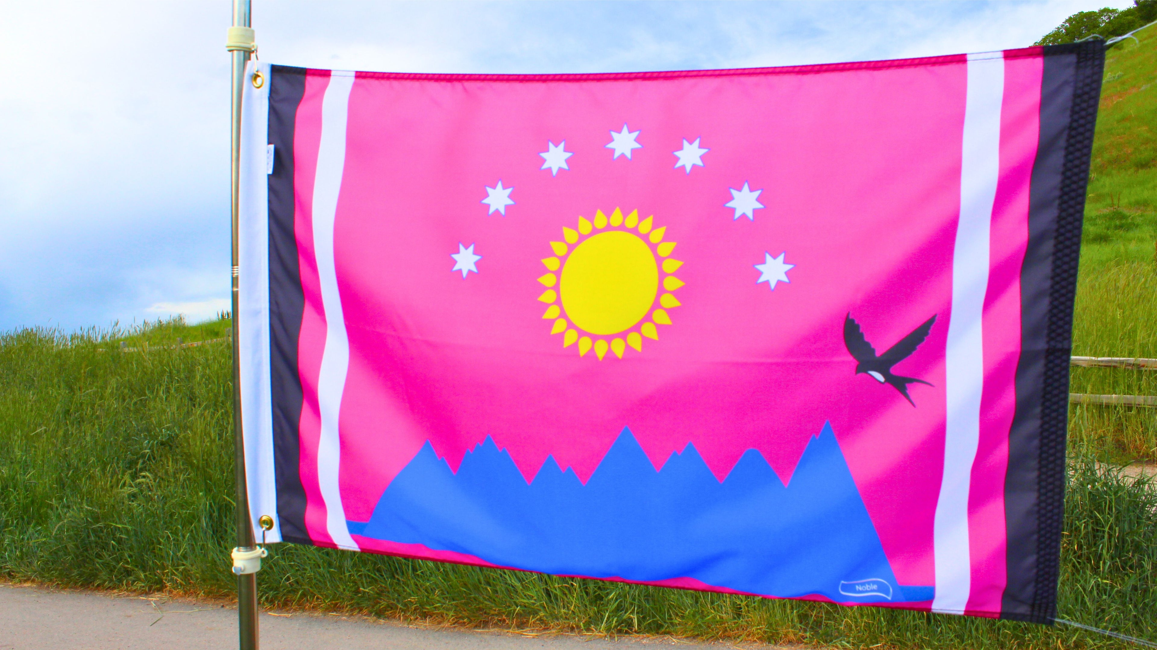 Flag with a sun, mountains, and a bird design on a pink background, waving in the wind.